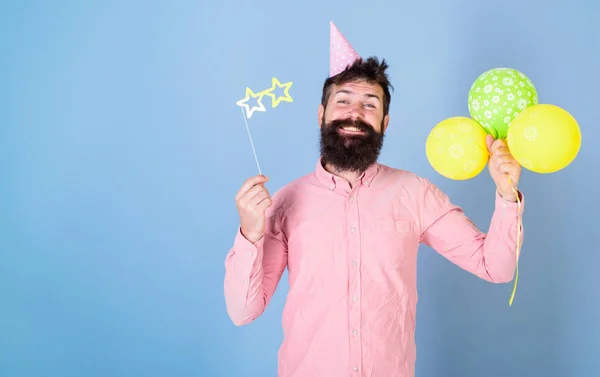 Hipster in sternförmiger Brille feiert Geburtstag. Parteizuschreibungen. Mann mit Bart im glücklichen Gesicht hält Luftballons, hellblauer Hintergrund. Mann mit Partyhut und Luftballons feiert — Stockfoto