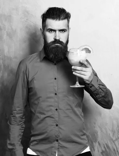 Bearded man, long beard. Brutal serious caucasian hipster with moustache in brown shirt holding tropical alcoholic fresh cocktail with orange piece on golden texture background