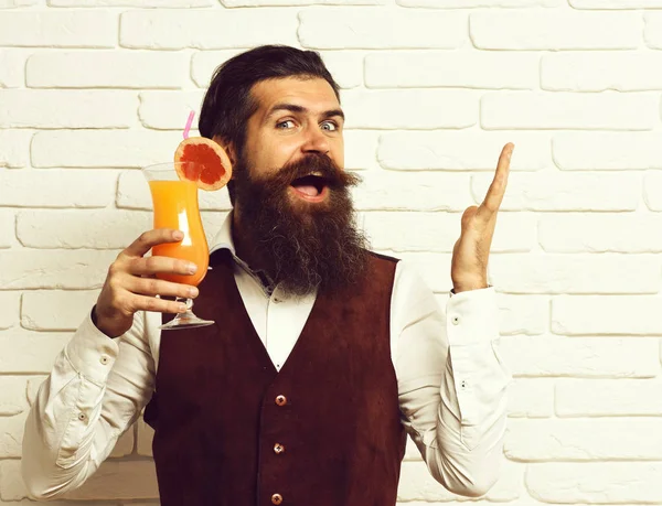 handsome bearded man with long beard has stylish hair on happy face holding glass of alcoholic beverage in vintage suede leather waistcoat on white brick wall studio background, copy space.