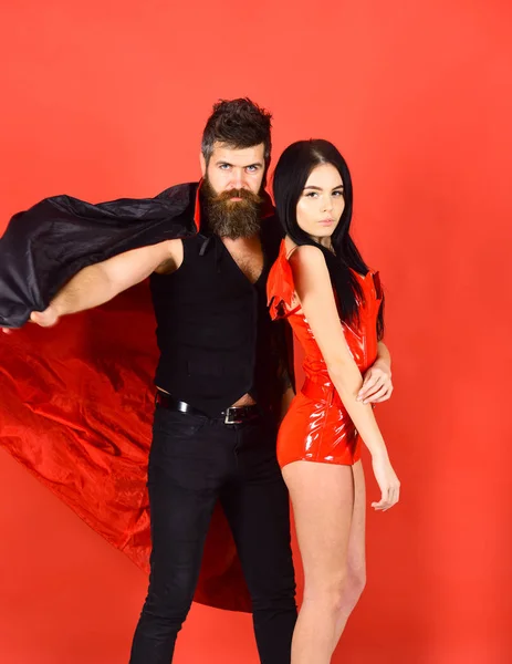 Attractive demon concept. Man and woman dressed like vampire, demon, red background. Couple in love, perfect match. Couple on strict face play role game. Vampire in cloak and sexy devil girl cuddling