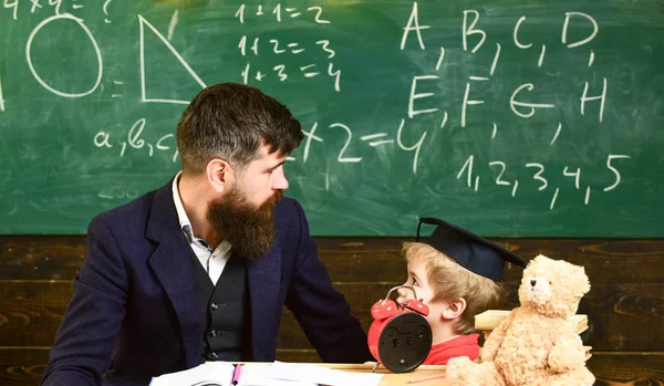 Rapaz, a criança na pós-graduação olha para rabiscos no quadro enquanto o professor explica. Professor com barba, o pai ensina o filho pequeno na sala de aula, quadro-negro no fundo. Conceito de lição individual — Fotografia de Stock