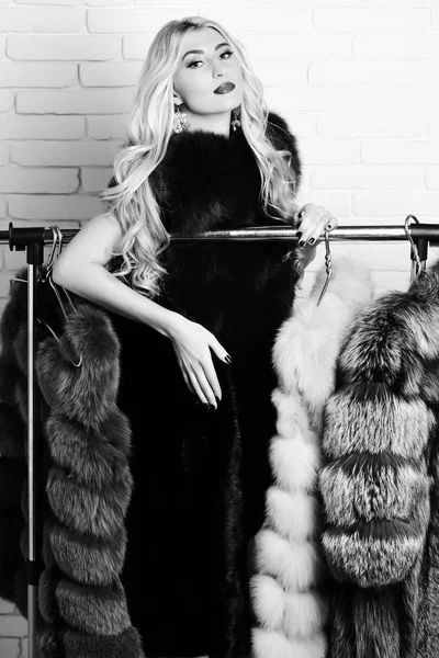 Jovem elegante sexy bonita mulher com belo longo cabelo loiro encaracolado no casaco de cintura de pele borgonha ou vermelha em pé sobre rack com cabides dourados no fundo estúdio parede de tijolo — Fotografia de Stock