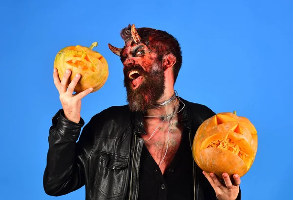 Man wearing scary makeup holds pumpkins on blue background.