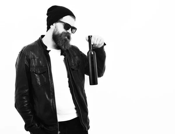 Bearded man, long beard. Brutal caucasian serious unshaven hipster holding bottle in black leather jacket, hat and glasses isolated on white studio background. — Stock Photo, Image