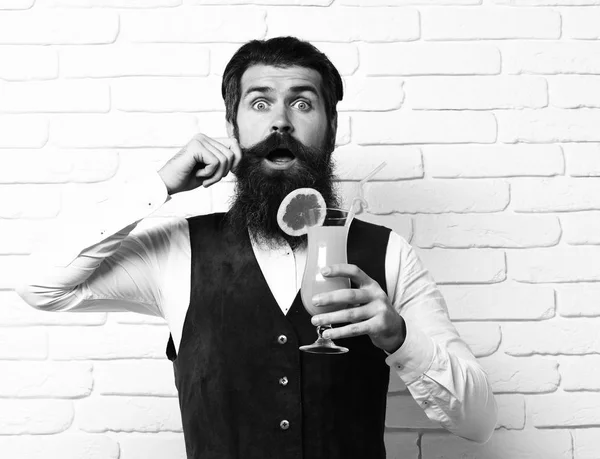 handsome bearded man with long beard and mustache has stylish hair on surprised face holding glass of alcoholic beverage in vintage suede leather waistcoat o white brick wall studio background