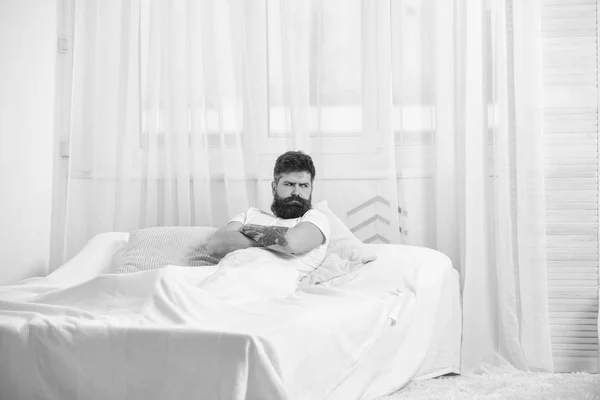 El tipo con cara seria e infeliz se ve enojado por la mañana. Hombre de camisa acostado en la cama, cortinas blancas en el fondo. Concepto de insomnio. Macho con barba cogida de la mano cruzada en el pecho, relajante — Foto de Stock