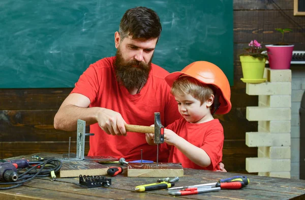 Отец с бородой учит маленького сына пользоваться инструментами, молотком, доской на заднем плане. Ребенок, занятый в защитном шлеме, учится пользоваться молотком вместе с отцом. Концепция отцовства — стоковое фото