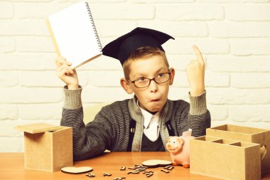 Genç sevimli öğrenci çocuk gri kazak ve beyaz tuğla duvar arka plan üzerinde sınıf mezuniyet şapkalı boşaltmak el ahşap numaraları pembe domuz domuz banka ile masa başında oturan gözlük