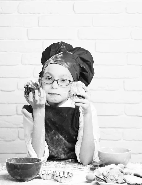 Young boy cute cook chef in red uniform and hat on stained face with glasses sitting on table with colorful bowls, tasty cookies, rolling pin and holding chocolate cakes on white brick wall background — Stock Photo, Image