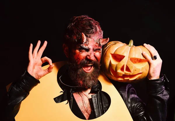Демон с улыбкой держит Джека Фонаря и показывает знак ОК. Концепция вечеринки на Хэллоуин. Человек в страшном макияже держит вырезанный тыквенный силуэт на шее — стоковое фото