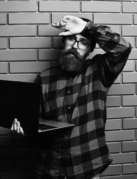 Hipster segurando laptop em vermelho camisa xadrez preto com chapéu e óculos no fundo do estúdio de parede de tijolo marrom. Barbudo brutal caucasiano hipster segurando laptop — Fotografia de Stock