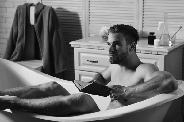 Concept de sexualité et de relaxation. Homme à la barbe et au visage attentionné lisant le livre. Guy lisant dans la salle de bain avec articles de toilette et chaise sur fond — Photo