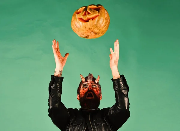 Man wearing scary makeup throws pumpkin up on green background. Halloween party concept. Devil or monster with October decorations.
