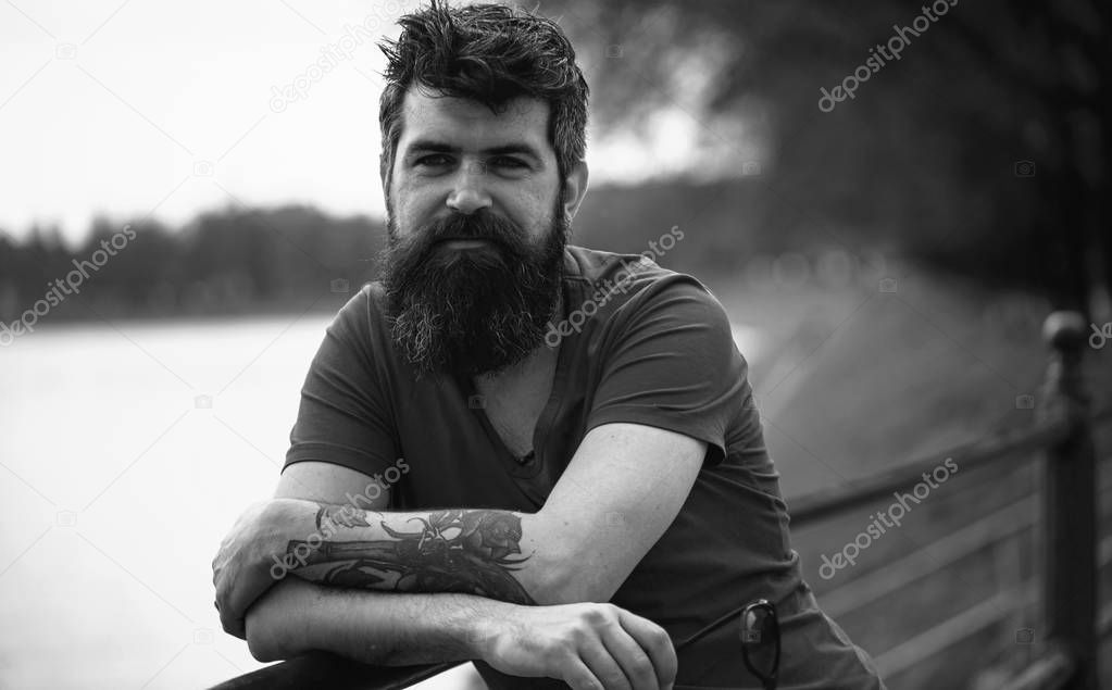 Man with beard and mustache with tattoo and sunglasses, riverside on background. Reflection and thoughts concept. Hipster on thoughtful face standing at riverside, thinking, dreaming, defocused.