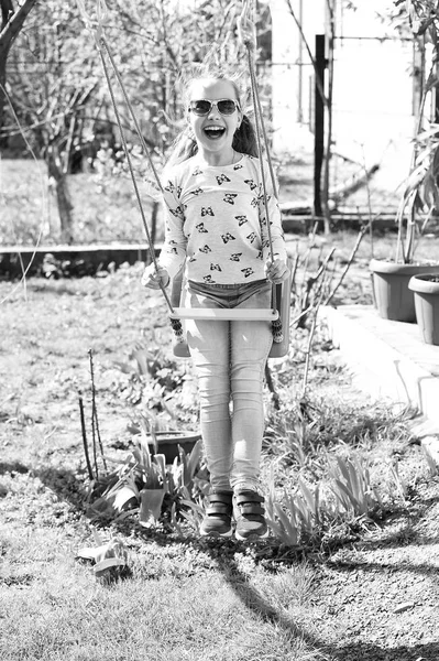 Χαρούμενη παιδική ιδέα. Ένα μικρό χαμόγελο στην κούνια στην αυλή του καλοκαιριού. Μόδα κορίτσι με γυαλιά ηλίου απολαύσετε αιώρηση την ηλιόλουστη μέρα. Ομορφόπαιδο χαμογελάει στην παιδική χαρά. Ελευθερία ξέγνοιαστος τρόπος ζωής και διασκέδαση — Φωτογραφία Αρχείου