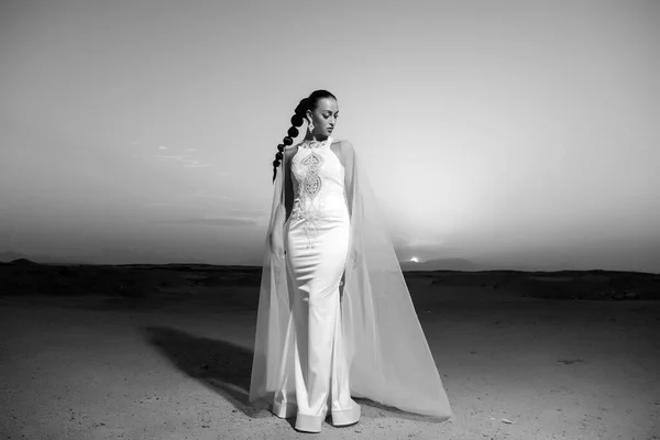 Sposa in abito da sposa sul cielo del tramonto. Donna vestita di bianco nel deserto. Donna sensuale con i capelli castani. Modella in dune di sabbia. Bellezza ragazza con look glamour e trucco — Foto Stock