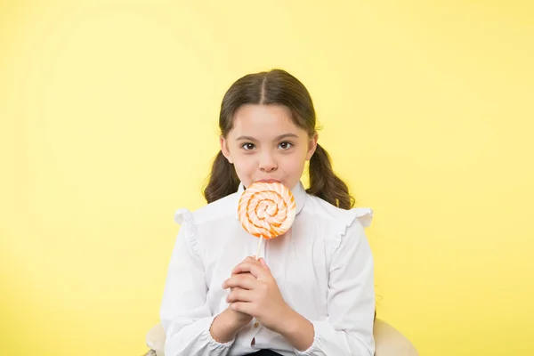 Unterernährung häufiges Problem. Mädchen Schüler Schuluniform mag Süßigkeiten Lollipop Süßigkeiten gelben Hintergrund. Mädchen niedlichen Kind Pferdeschwänze Frisur essen süßen Lutscher. Süßigkeiten in angemessenen Portionen ok — Stockfoto