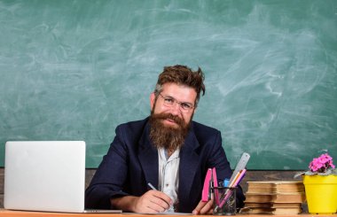 Evrak öğretmenler hayatın bir parçası. Ev ödevleri veya test kontrol öğretmen. Öğretmen Danışma dizüstü bilgisayar ile otur. Ödev kontrol edin. Öğretmen hipster gözlük sit sınıf kara tahta arka plan ile sakallı