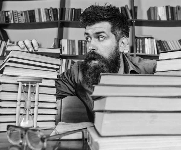 Concepto de fecha límite. Profesor o estudiante con barba se sienta en la mesa con gafas, desenfocado. Hombre en la cara conmocionada entre montones de libros, mientras estudiaba en la biblioteca, estanterías en el fondo . —  Fotos de Stock