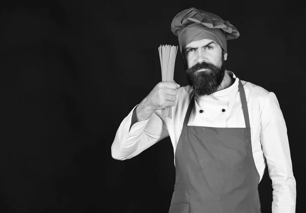 Chef-kok met bos van spaghetti. Man of hipster met baard houdt macaroni op zwarte achtergrond — Stockfoto