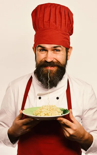 Man with beard and noodle in mouth holds tasty dish on white background. Restaurant cuisine concept. Cook with happy face