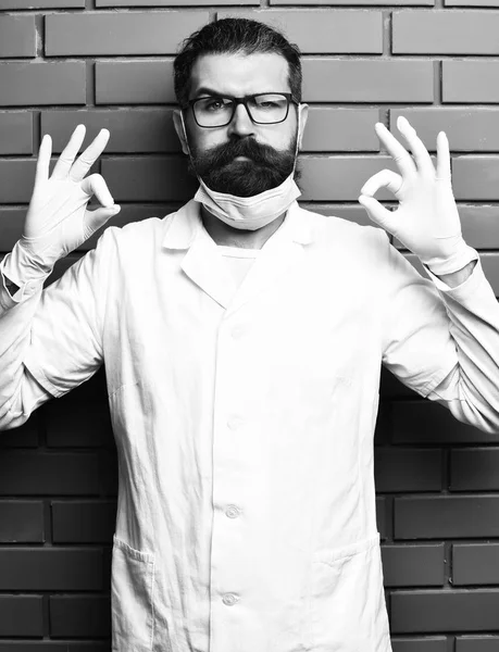 Μεταπτυχιακός φοιτητής στο ιατρικό εσθήτα, γυαλιά και γάντια σε καφετί τούβλο τοίχο studio φόντο. Ιατρική αντίληψη. Γενειοφόρος βάναυση Καυκάσιος γιατρό ή μεταπτυχιακός φοιτητής στο γάντια — Φωτογραφία Αρχείου