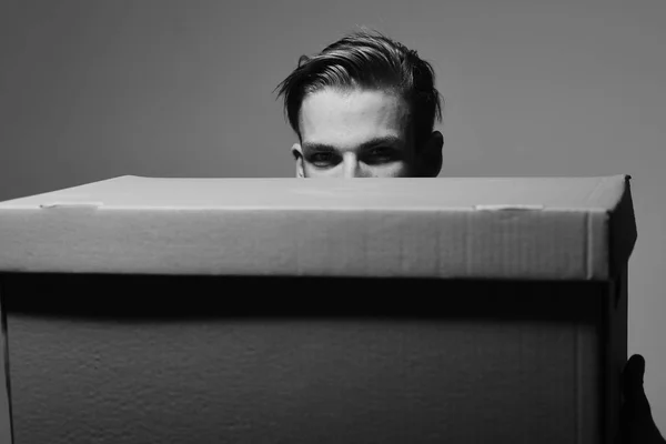 Loader holds box in front of face, close up. Delivery and moving concept. Man with messy hair