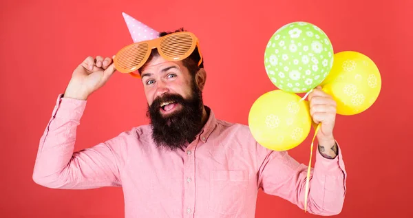 Hipster gucken aus einer riesigen Sonnenbrille. Mann mit Bart und Schnurrbart auf glücklichem Gesicht hält Bündel Luftballons, roter Hintergrund. Mann mit Partyhut und Luftballons feiert. Festkonzept — Stockfoto