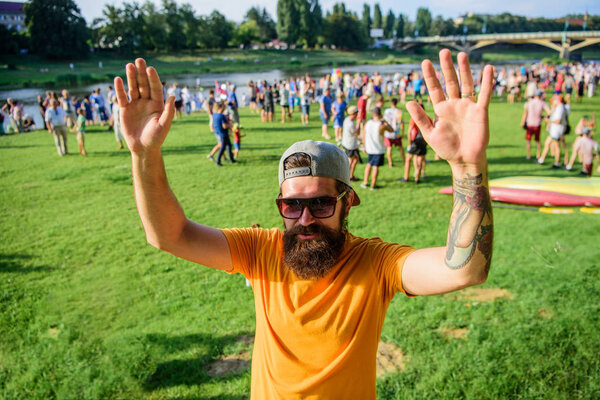 Хипстер в кепке рад встретить друга на пикнике или празднике. Рад с вами познакомиться. Человек бородатый хипстер перед толпой людей, машущих рукой зеленым фоном на берегу реки. Празднование городского события
