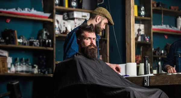 Um cliente Hipster a cortar o cabelo. Cliente com barba pronta para aparar ou arrumar. Processo de corte de cabelo conceito. Homem com barba coberta com capa preta esperando enquanto o barbeiro muda de categoria clipper — Fotografia de Stock