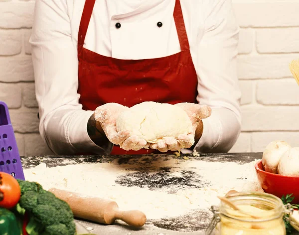 Férfi kezében tartsa gyúrt tészta, a fehér téglafal háttér, közelről. Szakács teszi a tésztát. Cook, Burgundia egységes ül tábla zöldségekkel — Stock Fotó