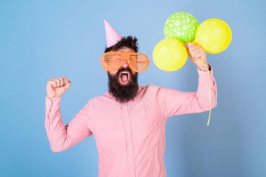 Hipster dev gözlük içinde Doğum günü kutluyor. Kutlama kavramı. Hava balonları ile parti şapkalı adamı kutluyor. Mutlu yüz tutun hava balonları, açık mavi renkli sakallı