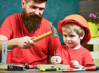 Boy, child busy in protective helmet learning to use hammer with dad. Father with beard teaching little son to use tools, hammering, chalkboard on background. Handyman concept clipart
