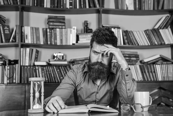 Hombre en el libro de lectura de la cara concentrada, estudio, estanterías en el fondo. Concepto de autoeducación. El hombre se sienta a la mesa con taza y reloj de arena. Profesor o estudiante con barba estudiando en la biblioteca —  Fotos de Stock