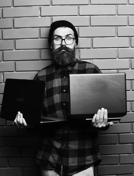 Bearded man, long beard. Brutal caucasian surprised unshaven hipster holding laptops in red black checkered shirt with hat and glasses on brown brick wall studio background.