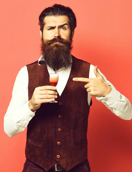 Handsome bearded man with long beard and mustache on serious face holding glass of alcoholic shot in vintage suede leather waistcoat on red studio background. — Stock Photo, Image