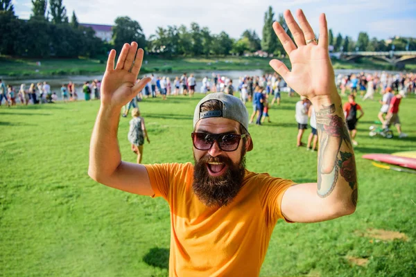 Happy to meet you. Man bearded hipster in front of crowd people waving hand green riverside background. Urban event celebration. Hipster in cap happy to meet friend at event picnic fest or festival