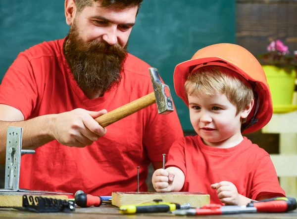 Boy, child busy in protective helmet learning to use hammer with dad. Father with beard teaching little son to use tools, hammering, chalkboard on background. Handyman concept