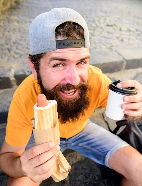 Hipster comer cachorro-quente enquanto se sentar em escadas ao ar livre. Hipster desfrutar de cachorro quente e beber xícara de papel. Homem barbudo desfrutar de comida de rua fundo urbano. Faça uma pausa para lanchar. Conceito de cultura alimentar urbana — Fotografia de Stock