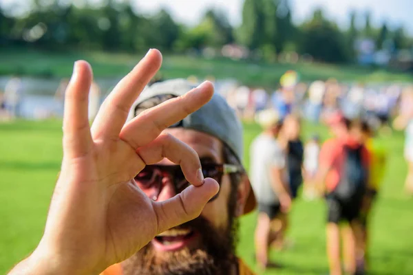 Man bearded in front of crowd riverside background. Book ticket summer festival in advance. Hipster visiting fest festival. Man cheerful face looks through ok gesture. Be sure in your summer vacation — Stock Photo, Image