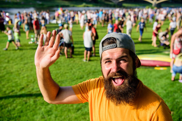Хипстер в кепке посещает пикник или фестиваль. Празднование городского события. Мужчина машет рукой солнечный день на улице. Человек бородатый хипстер перед толпой людей зеленый фон поля
