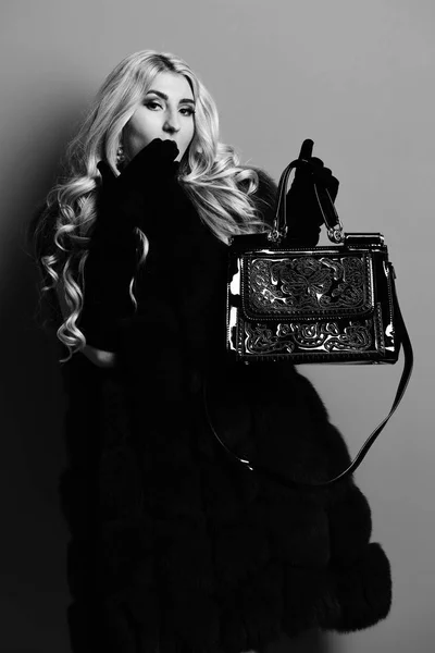 young fashionable sexy pretty woman with beautiful long curly blonde hair in waist coat of burgundy fur and black velvet gloves showing white fashion bag on red studio background