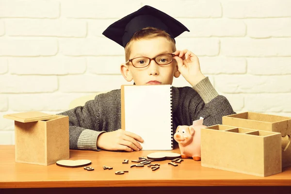 Ung söt elev pojke i grå tröja och glasögon sitter vid skrivbordet med copybook i handen trä siffror rosa gris bank i examen mössa i klassrummet på vit tegelvägg bakgrund, kopiera utrymme. — Stockfoto