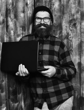 Sakallı adam, uzun sakallı. Kahverengi, eski, ahşap stüdyo arka planında şapka ve gözlüklerle kırmızı kareli tişörtlü, mutlu, traşsız hippi gülümsemesi..