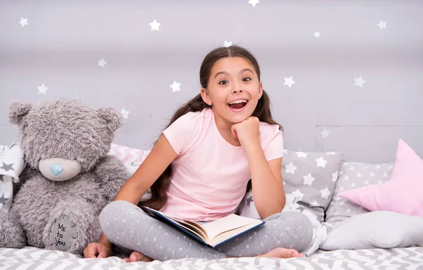 Guess her favorite fairy tale. Girl child sit bed with teddy bear read book. Kid prepare to go to bed. Pleasant time in cozy bedroom. Girl kid long hair cute pajamas relax and read book to bear toy