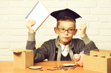 Gri süveterli, gözlüklü, elinde defteriyle masada oturan yakışıklı öğrenci. Ahşap numaralar. Mezuniyet şapkasında pembe domuzcuk bankası.