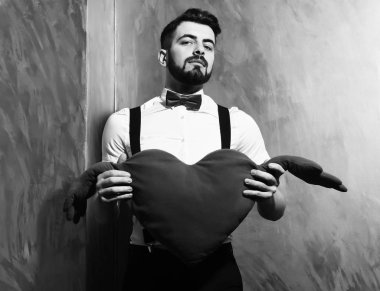 Sakallı adam elinde kırmızı pelüş bir kalp ve ciddi bir yüz tutuyor.