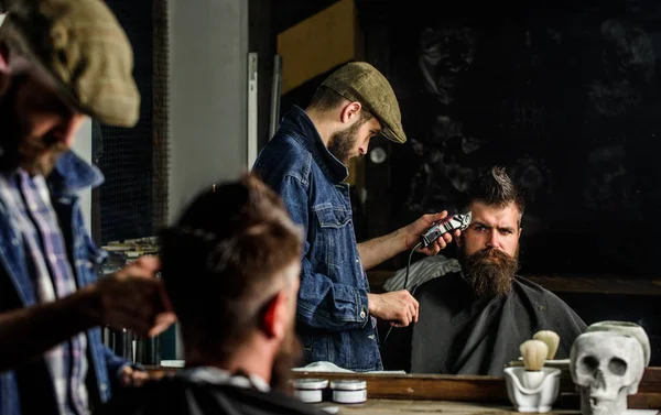 Haircut concept. Barber with hair clipper works on hairstyle for man with beard, barbershop background. Barber styling hair of brutal bearded client with clipper. Hipster client getting haircut