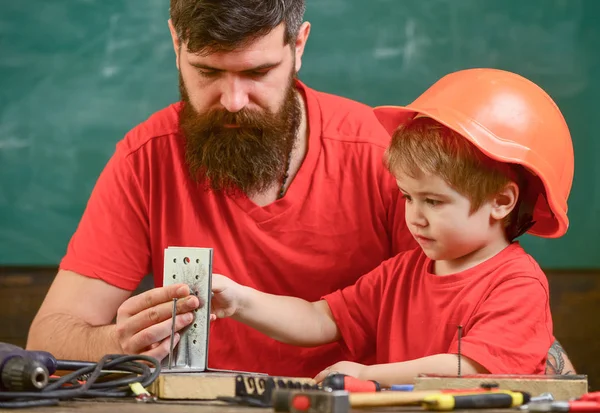 少年は、保護用のヘルメットで忙しい子になります、手でお父さんと修復します。ほとんどアシスタントのコンセプトです。髭のツールを使用して、背景に黒板の教室で少し息子を教える父 — ストック写真