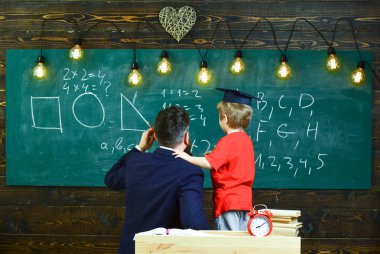 Preparation for school concept. Teacher with beard, father teaches little son in classroom, chalkboard on background. Boy, child in graduate cap listening teacher, chalkboard on background, rear view clipart